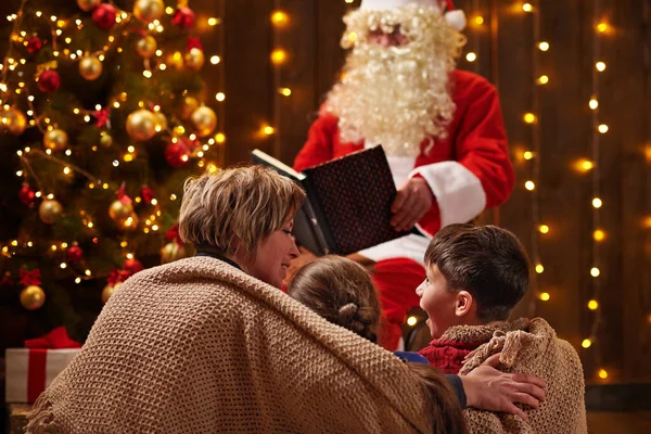 Санта Клаус читає книжку для сім'ї. Мати з дітьми сидять у приміщенні біля прикрашеного ксам дерева з сяйвом - Веселого Різдва і Веселих свят.! — стокове фото