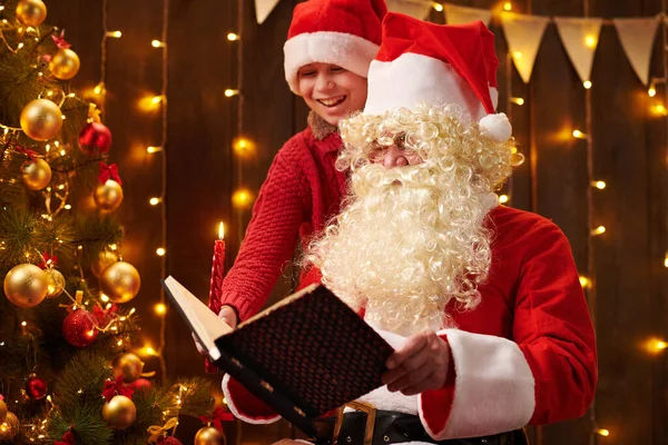 Père Noël et santa aide garçon livre de lecture, assis à l'intérieur près décoré arbre de Noël avec des lumières Joyeux Noël et Joyeuses Fêtes ! — Photo