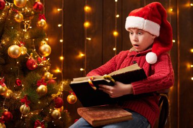 Genç çocuk kitap okuyor, kapalı bir yerde Noel Baba yardımcısı gibi giyinmiş, ışıklarla süslenmiş Xmas ağacının yanında oturuyor.!