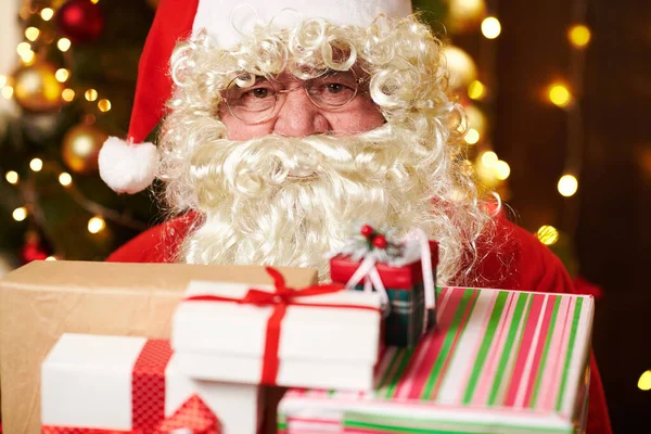 サンタクロースは多くの贈り物でポーズをとって、照明付きの装飾されたクリスマスツリーの近くに屋内に座っています-メリークリスマスとハッピーホリデー! — ストック写真