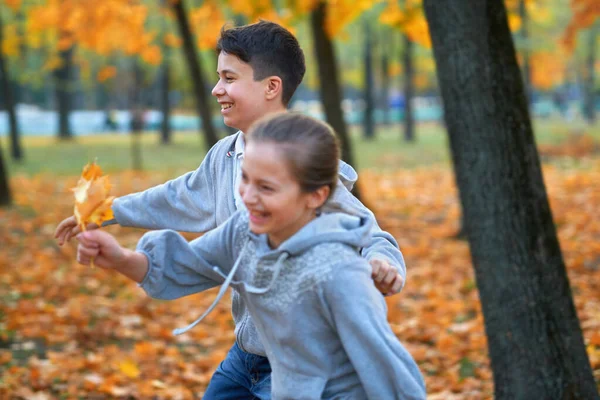 Девочка и мальчик-подросток отдыхают в осеннем городском парке, бегают, улыбаются, играют и веселятся. Ярко-желтые деревья и листья — стоковое фото