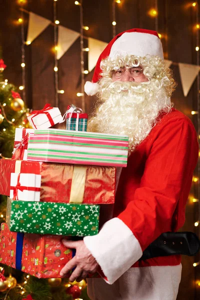 Père Noël posant avec de nombreux cadeaux, assis à l'intérieur près de l'arbre de Noël décoré avec des lumières Joyeux Noël et Joyeuses Fêtes ! — Photo