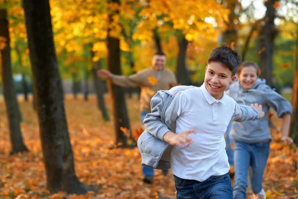 Szczęśliwy rodzina mający święto w jesienny miejski park. Dzieci i rodzice biegają, uśmiechają się, bawią i bawią. Jasnożółte drzewa i liście — Zdjęcie stockowe