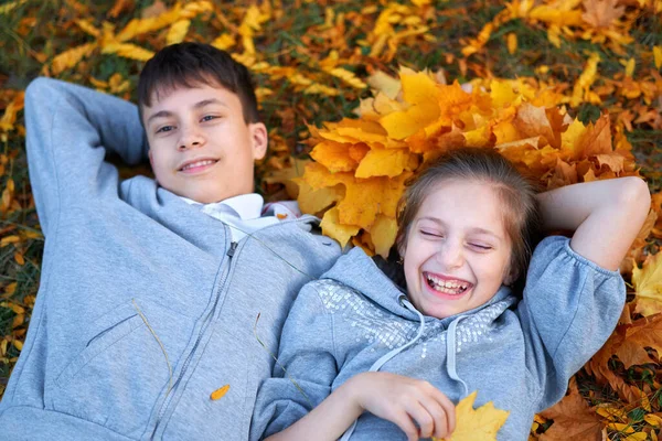 Niña y niño adolescente teniendo vacaciones en el parque de la ciudad de otoño, acostado en la hierba, posando, sonriendo, jugando y divirtiéndose. Árboles y hojas de color amarillo brillante —  Fotos de Stock