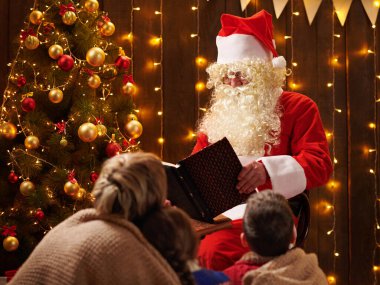 Noel Baba aile için kitap okuyor. Anne ve çocuklar dekore edilmiş Noel ağacının yanında ışıklarla oturuyorlar Mutlu Noeller ve mutlu tatiller!