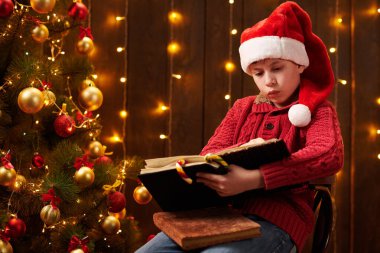 Genç çocuk kitap okuyor, kapalı bir yerde Noel Baba yardımcısı gibi giyinmiş, ışıklarla süslenmiş Xmas ağacının yanında oturuyor.!