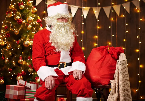 Père Noël posant avec un sac de cadeaux, assis à l'intérieur près décoré arbre de Noël avec des lumières Joyeux Noël et Joyeuses Fêtes ! — Photo