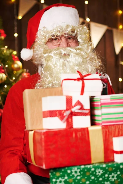 サンタクロースは多くの贈り物でポーズをとって、照明付きの装飾されたクリスマスツリーの近くに屋内に座っています-メリークリスマスとハッピーホリデー! — ストック写真
