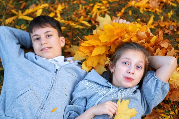 Niña y niño adolescente teniendo vacaciones en el parque de la ciudad de otoño, acostado en la hierba, posando, sonriendo, jugando y divirtiéndose. Árboles y hojas de color amarillo brillante —  Fotos de Stock