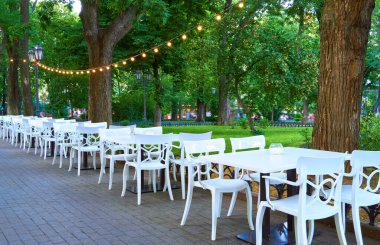 Açık hava sokak kafesi - beyaz sandalyeler ve masalar, aydınlatmayla süslenmiş, şehir parkı ve yürüyen insanlar