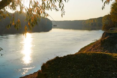 Günbatımında renkli sonbahar ormanı manzarası, nehir ve gökyüzü yakınındaki ağaçlar, suya yansıyan parlak güneş ışığı.