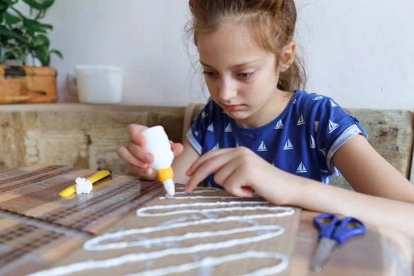 女の子は工芸品や接着剤の段ボールを作って家のキッチンに座り — ストック写真