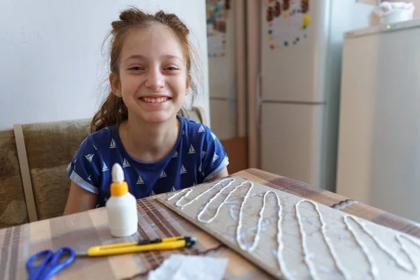 女の子は工芸品や接着剤の段ボールを作って家のキッチンに座り — ストック写真