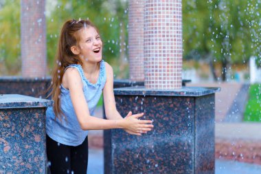 Şehir parkındaki çardakta düşen su damlalarıyla oynayan bir kız, aydınlık bir yaz günü.