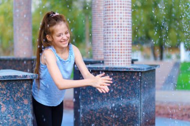 Şehir parkındaki çardakta düşen su damlalarıyla oynayan bir kız, aydınlık bir yaz günü.