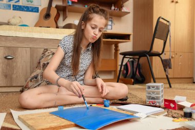 Kız evde resim çiziyor, sanat eseri yapıyor, kağıt, boya ve fırçalardan yaratıcı sanat eserleri yapıyor.