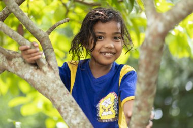 BANDA NEIRA, MALUKU ISLANDS, INDONESIA, DECEMBER 17, 2017 : Portrait of a smiling sweet little girl climbing at a tree in Banda Neira, Maluku islands, Indonesia. clipart