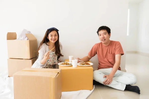 一对亚洲情侣的肖像坐在地板上 吃着烤鸭蛋面条在纸板搬运盒在新房 建筑与房地产业开创新的家庭生活 图库照片