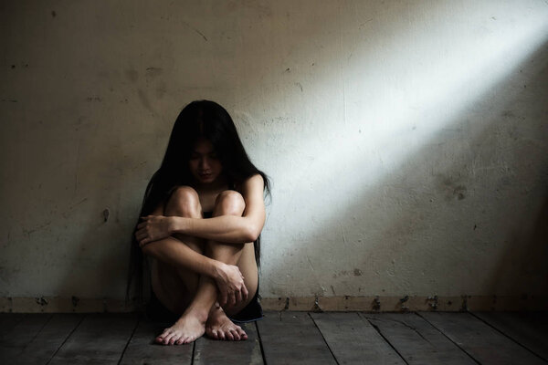 Stressed beautiful Asian woman sitting on wood floor with depress feeling. Sexy prostitute girl sad in enpty dark room with light shade by the window.
