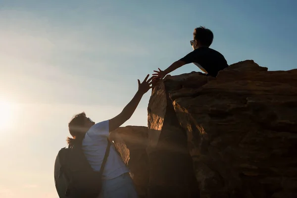 hiking woman climbing up to hill summit  while son try to grab hand during sunset. Healthy mother hike to mountain cliff peak. Family activity and outdoor extreme sport concept.