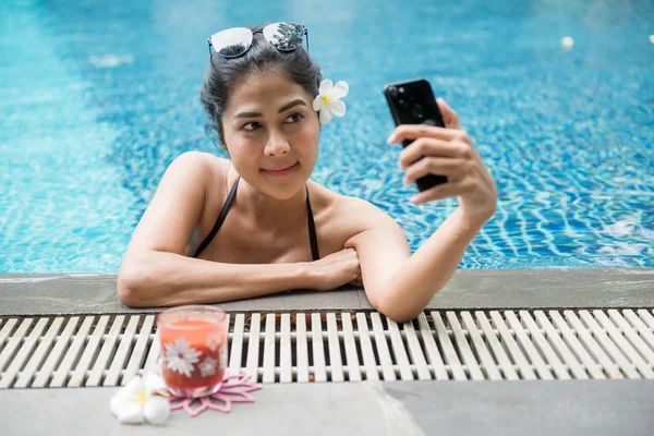 迷人的性感亚洲妇女与黑色两件泳装和太阳镜自拍自己的智能手机 Nearat 游泳池边 美丽的女孩在暑假放松 图库图片