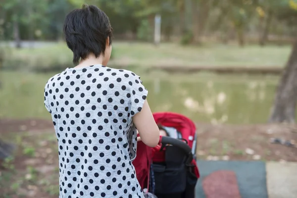 Mother wheel red stroller to let her child daugther to relax in summer park. Mom take care of baby gir in garden. Motherhood or Parent in duty.