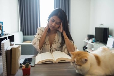 Stresli güzel Asyalı kadın ile şirin egzotik stenografi kedi tabloda metin kitap okuyun. Sevimli evcil hayvan ile evde kadın çalışma çalışma. kız baş ağrısı final sınavı için okuma.