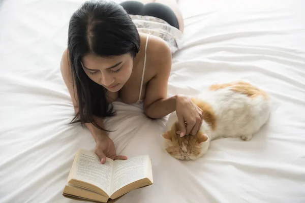 Sexy Asian tan woman read fiction text book on white bed while cute exotic shorthair cat sleep beside with copy space for text. Relax lifestyle in bedroom during weekend.