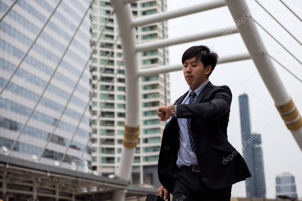Anxious Asian businessman run and look at watch to check time on city walk in rush hour. Young man late for train transportation, work, meeting. Office life and business competition.