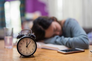 Yorgun öğrenci kadın uyku metin kitap içinde ev ile portre çalar saat. Ciddi exhauasted Asyalı kız wok final sınavı için hazırlamak için kitap okuyarak zor.