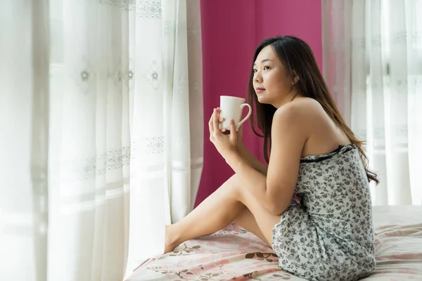 Asian Chinese woman drink hot coffee in the morning near window bedroom. Refreshment to start new day.