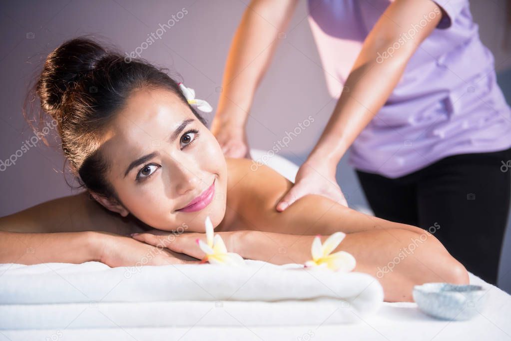 portrait of attractive Asian beautiful woman in spa club. Body care treatment by Thai oil. Cute girl having massage at back by professional massager hands with candle and white Plumeria on bed.