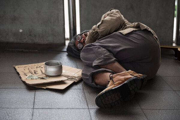 Old sick beggar or Homeless dirty man sleep on footpath with donate bowl, dollar bill, coin, paper cardboard with help text. feeling cold during winter with bad weather.
