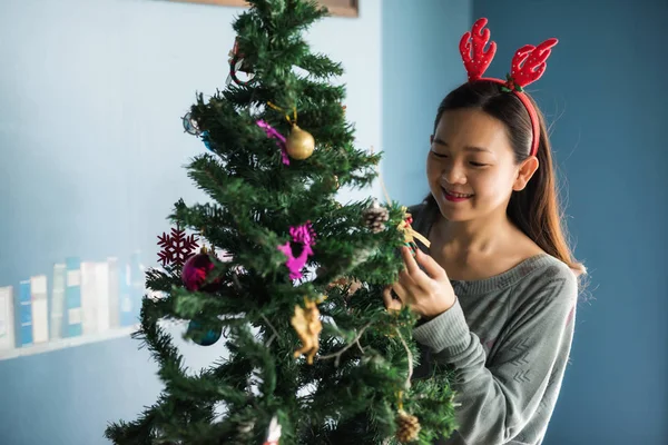 Asian Chinese happy girl with reindeer costume decorate gifts on Xmas tree. Attractive cute woman celebrate Christmas holiday in house with copy space for text.