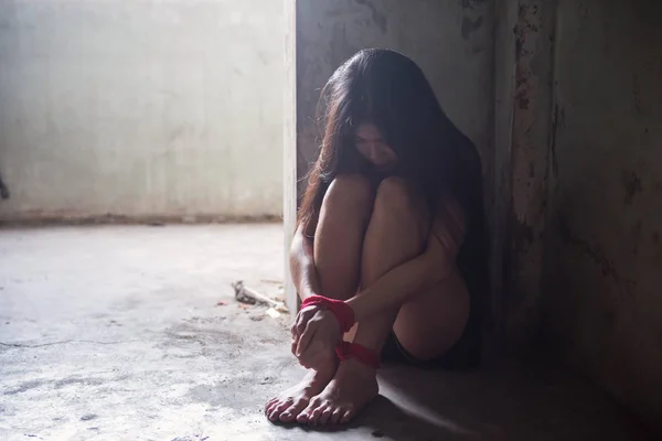 落ち込んで若いアジアの人質の女性は 汚い木製の床の上に座って赤いロープで手を縛っていた 放棄暗い部屋の窓から光の縁を持つ悲しい少女 菜種と犯罪 社会問題のコンセプト — ストック写真