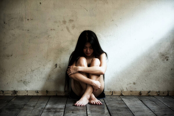 Depress beautiful Asian woman sitting on wood floor with depress feeling. Sexy prostitute girl sad in empty dark room with light shade by the window.