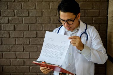 Asian doctor, 40-50 years old, with white lab coat reading patient record report on paper to analyst symptom in hospital. Healthcare and Medical concept clipart