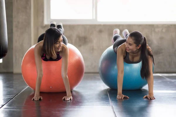 Happy smiling sporty lesbian LGBT couple or female friends in plank position exercising on fitness balls in gym. Women working out together for bodybuilding. Teamwork and achievement concept