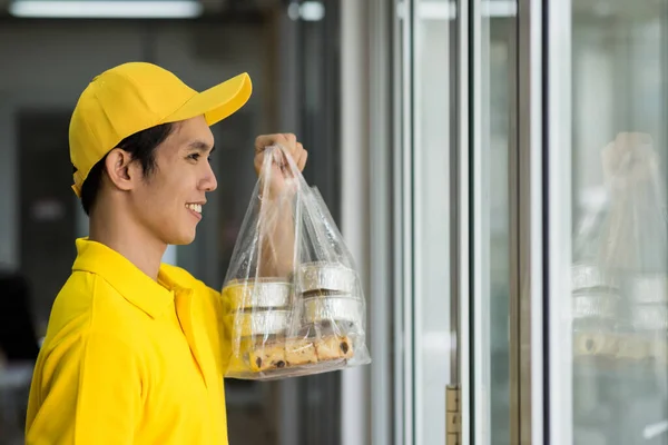 Smiling delivery man in yellow uniform delivering cake and snack dessert in plastic bags in office building. Handsome Asian young deliveryman with service mind carry and raise food. New normal concept