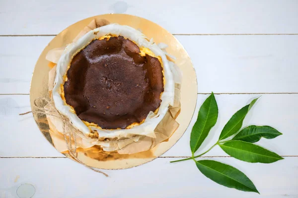 Basque burnt cheesecake wrapped by Parchment Paper on white wood paint with natural green leaf by aerial top view. Trendy homemade Baked bakery dessert and supplementary career in new normal.