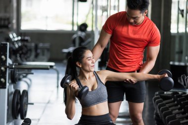 Spor salonundaki Asyalı güzel bir kadına kol pozisyonu değiştirerek Dumbbell egzersizi yaptıran kişisel antrenör. Vücut geliştirme ve sağlıklı yaşam için sandalyede çalışan mutlu bir kız..