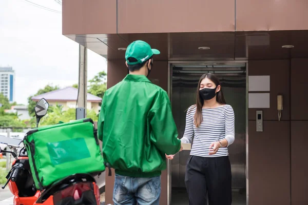 courier delivery man with face mask and green jacket uniform deliver pizza food box to female customer in front of condo or apartment near motorbike. covid19  with new normal business by online app.