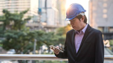 Kore 'li kıdemli mühendis, 40' lı yaşlarda, kasklı, akıllı telefon kullanarak proje planlarını ve gün batımında inşaat alanında zaman çizelgesini kontrol ediyor. İş afişi arkaplanı.