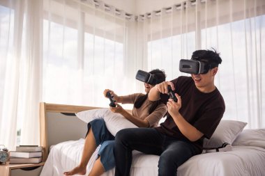 Yataktaki heyecanlı Asyalı çiftin portresi sanal gerçeklik gözlüğü ve evdeki kablosuz joystick ile oyun konsolu oynuyor. Hafta sonları iyi eğlenceler ve mutlu yaşam tarzları. Covid19 sırasında evde kal..