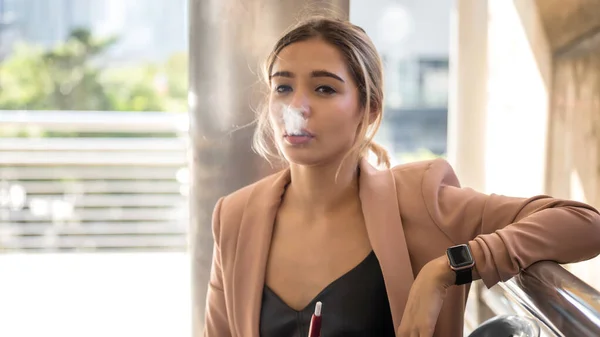 Beautiful American Businesswoman smoking an e-cigarette. Attractive girl smoke during lunch break outside office building in modern city.