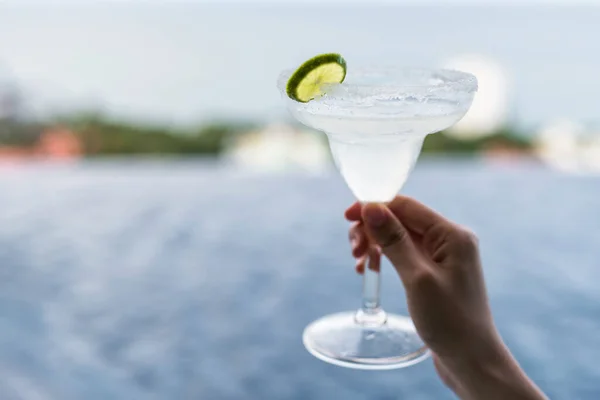 Frozen margarita cocktail with slice of lime in glass on woman hands in front of rooftop swimming pool with sea background.
