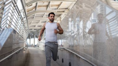 Yakışıklı Latin Amerikalı sakallı spor elbiseli genç adam şehir ya da şehirdeki skywalk köprüsünde koşuyor. Vücut geliştirme ve sağlıklı yaşam tarzı konsepti. Kasabada koşan sporcu çocuk..