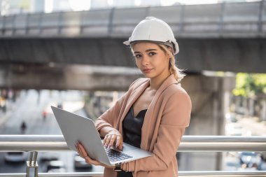 Yüzünde şapkası ve ceketi olan gülümseyen Amerikalı mühendis kadının portresi şehir merkezinde akşam ışığında dizüstü bilgisayarla çalışıyor. Çekici iş kadını proje planını kontrol et ya da İnternet açık artırmasıyla teklif ver.