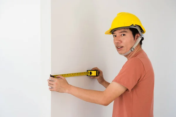 Asian Interior engineer man with yellow hardhat using measuring tape to measure empty wall of new house. Furniture or cabinet built in for modern blank home. Real estate industry.