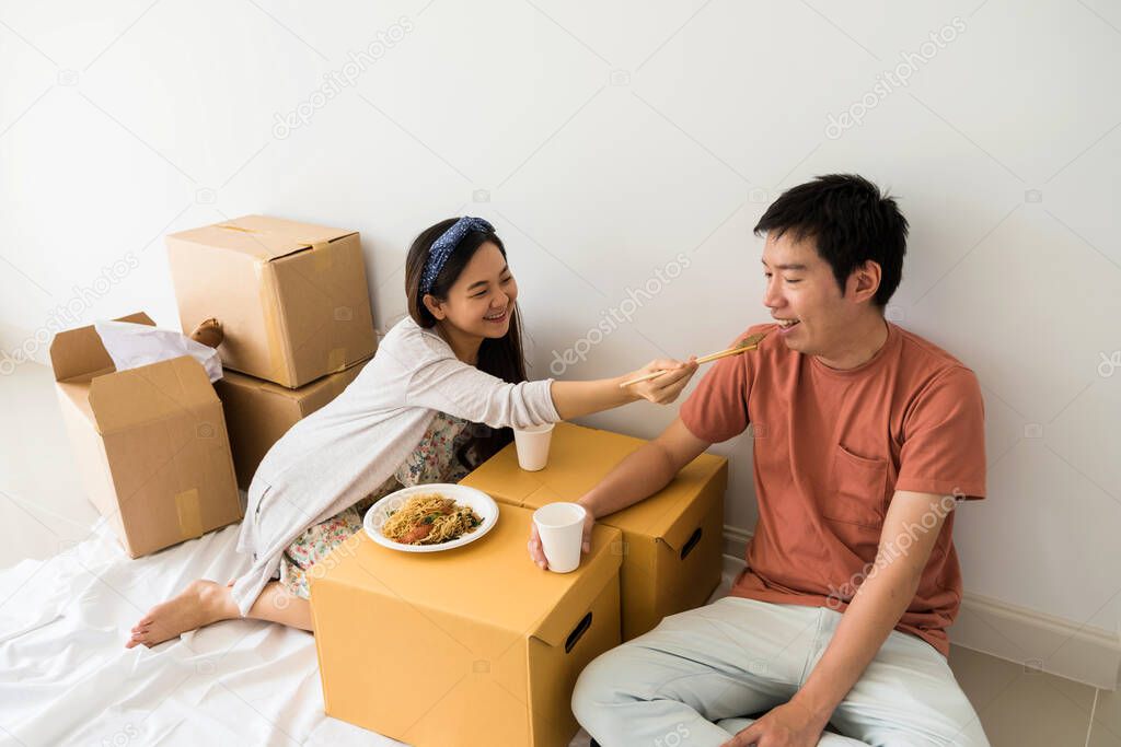 Young Asian couple sit on floor and eat roasted duck egg noodle on cardboard moving boxes to be unpacked stuff at new house. Home mortgage loan and Real estate to start new family life.